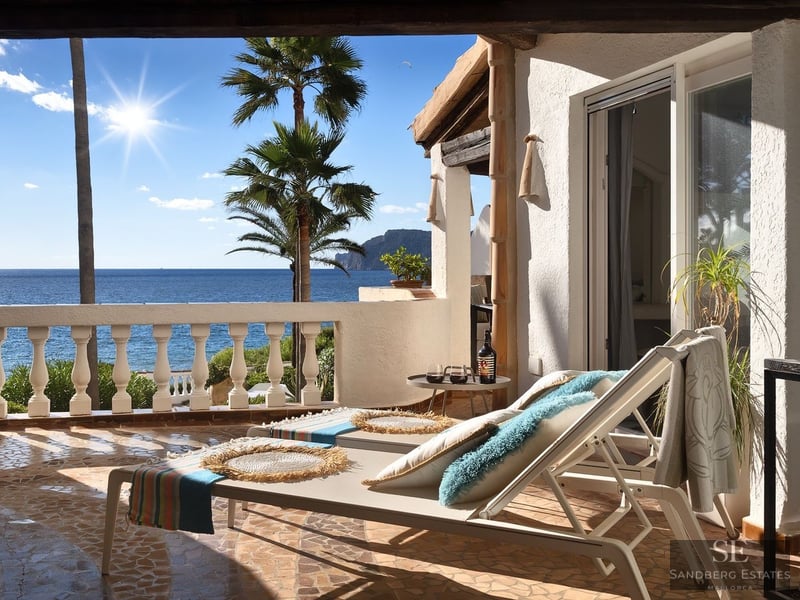 Tumbonas blancas en una terraza mediterránea con vistas al mar y palmeras bajo un sol radiante.