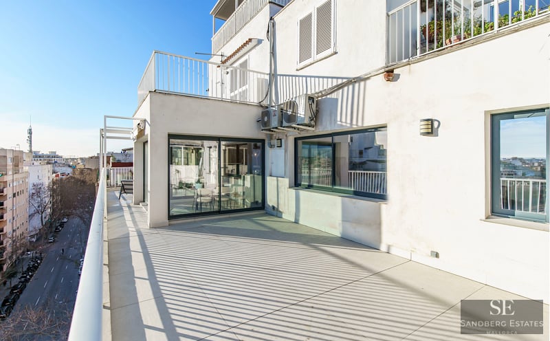 Sunny terrace with light tiling, white railings, and a view of a city street under a clear blue sky.
