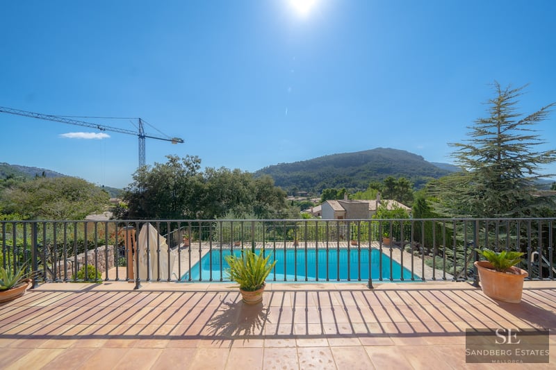 View from a terrace with iron railing overlooking a blue swimming pool and green mountains under a bright sun.