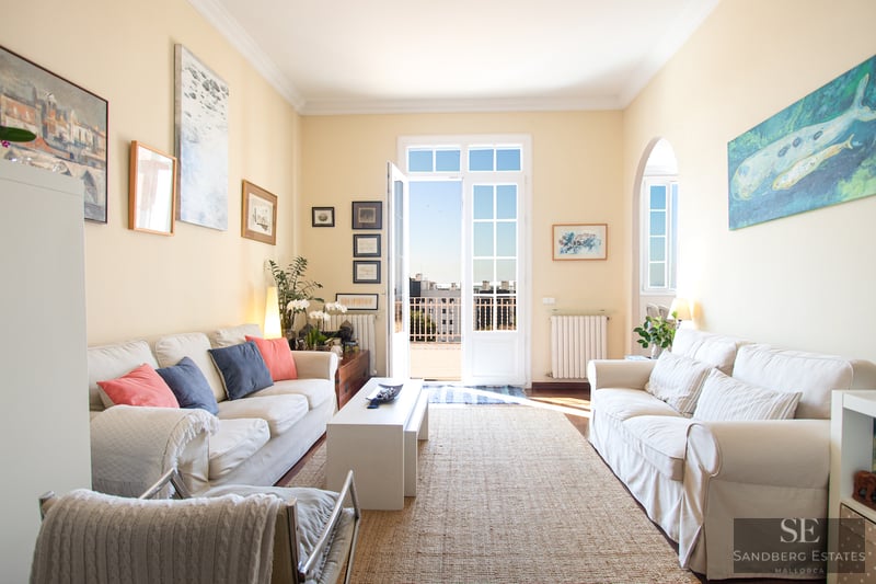 Sonnendurchflutetes Wohnzimmer mit weißen Sofas, Juteteppich und Fenstertüren, die auf einen Balkon mit Stadtblick führen.