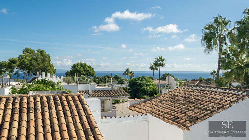 Elevated view of terracotta roofs, white walls, and palm trees with the blue sea in the background under a sunny sky.
