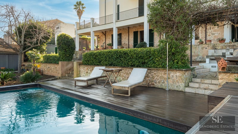 Turquoise swimming pool with dark wood deck, sun loungers, and a rustic stone villa in the background.
