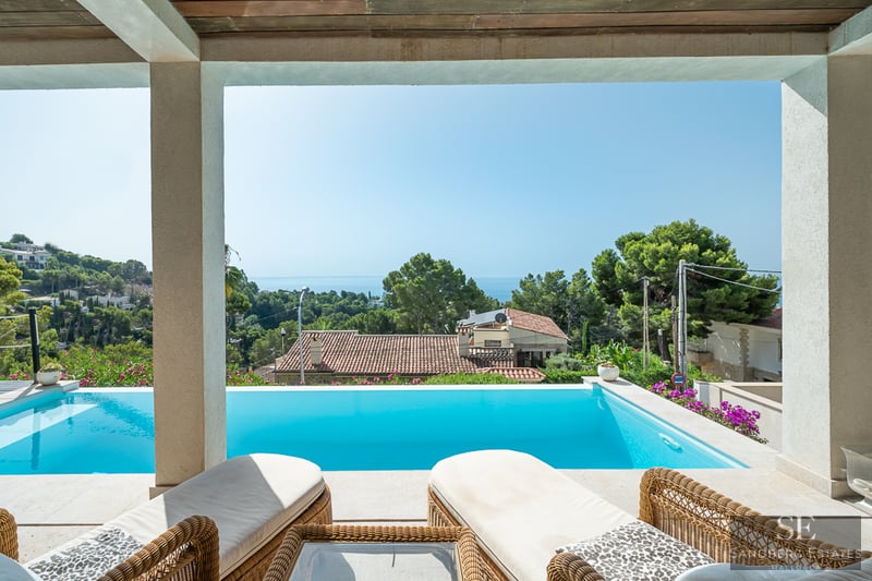 Blick von einer schattigen Terrasse über einen Infinity-Pool auf das Mittelmeer und üppige grüne Hügel.