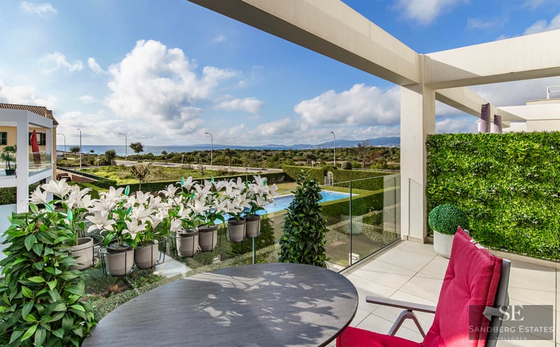 Luxury terrace with a round table, red chair, and glass railing overlooking a garden, pool, and the sea.
