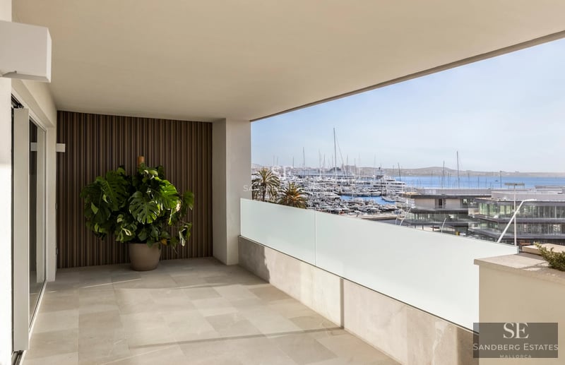 Spacious balcony with sea view. Glass railing, wooden floor, modern furniture, and lush vegetation.