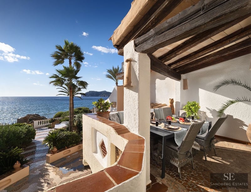 Terraza techada con vigas de madera, mesa de comedor, sillas de mimbre y vistas espectaculares al mar y palmeras.