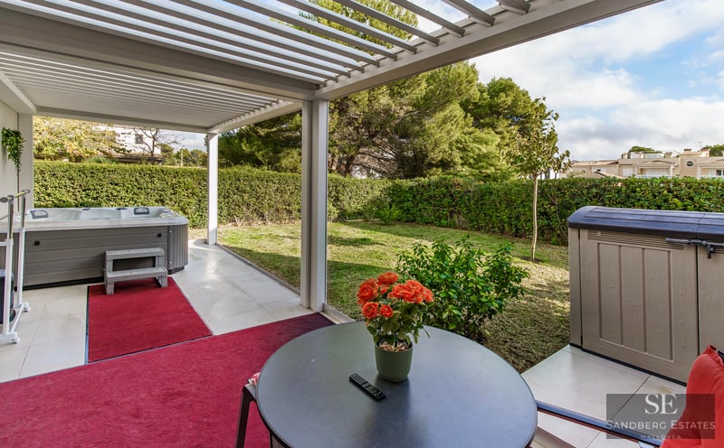 Outdoor terrace with a white pergola, grey hot tub, red rugs, and a view of a green garden with hedges.