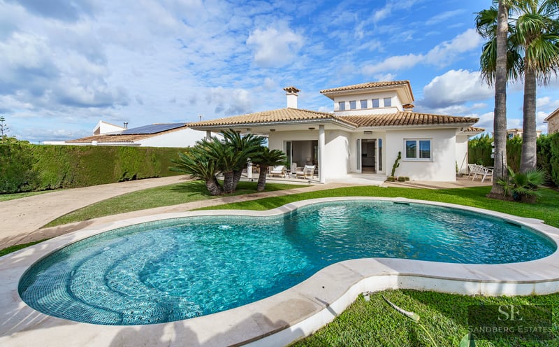A turquoise kidney-shaped swimming pool surrounded by green grass and palm trees in front of a Mediterranean white villa.