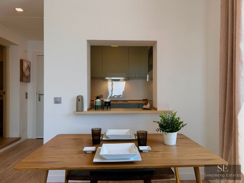 Wooden dining table set for two in front of a wall opening looking into a modern kitchen.