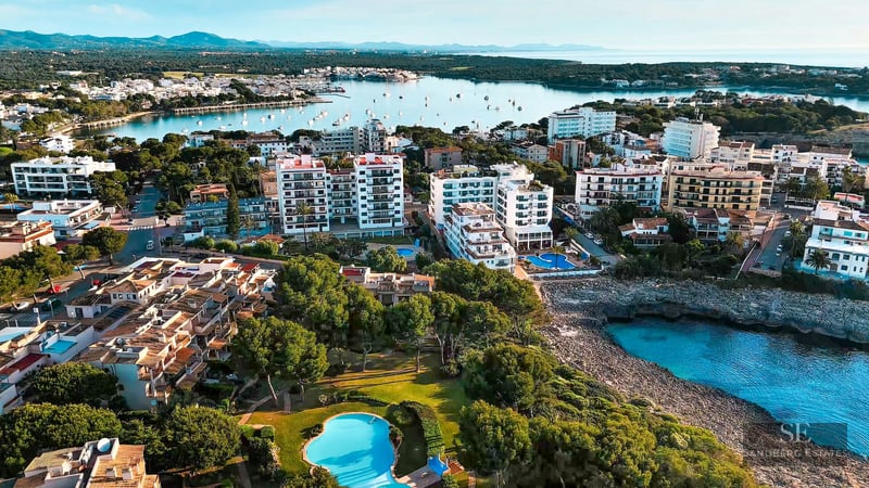 Aerial drone shot showing white buildings, swimming pools, and turquoise water coves surrounded by lush greenery.