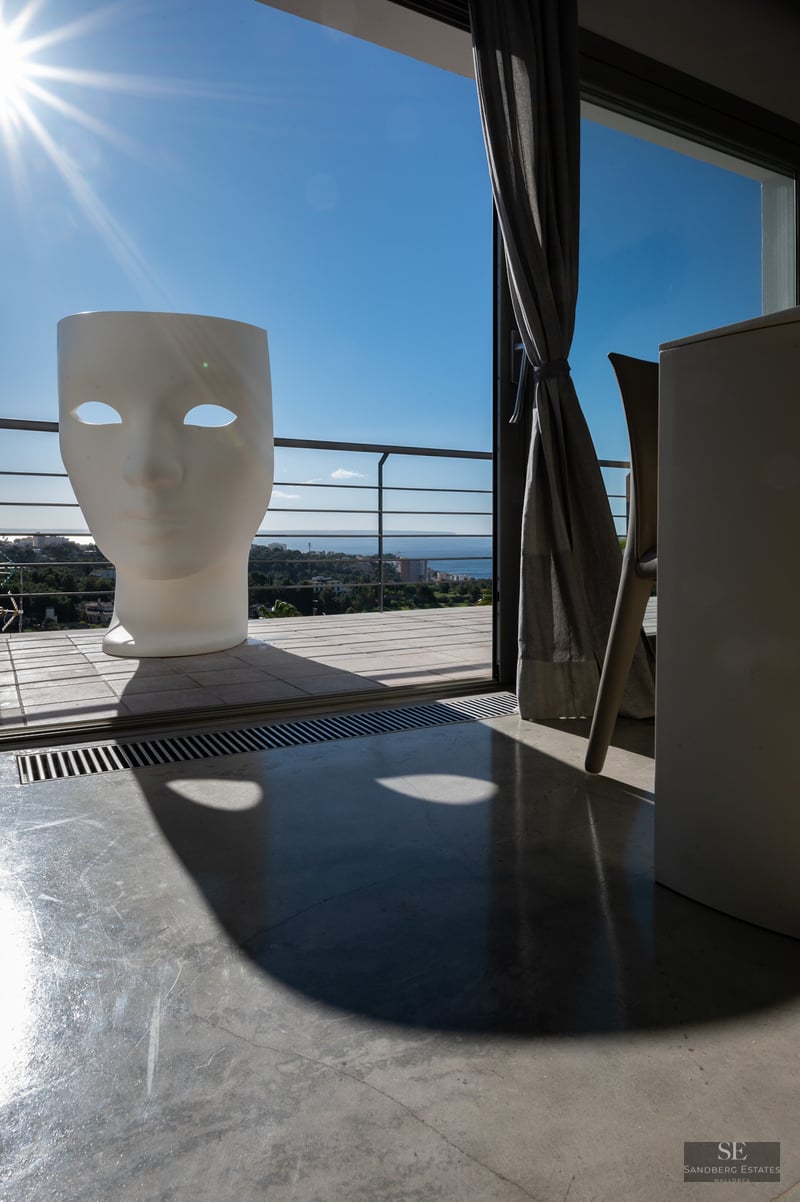 Interior view looking out to a sunny terrace featuring a giant white face-shaped chair and panoramic sea views.
