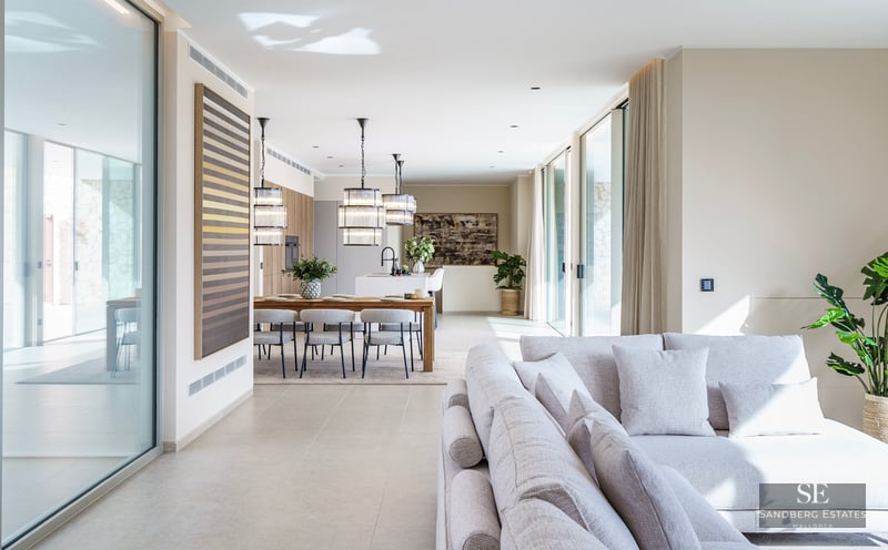 Spacious modern living room with light gray sofa, wooden dining table, and designer pendant lights by large glass doors.