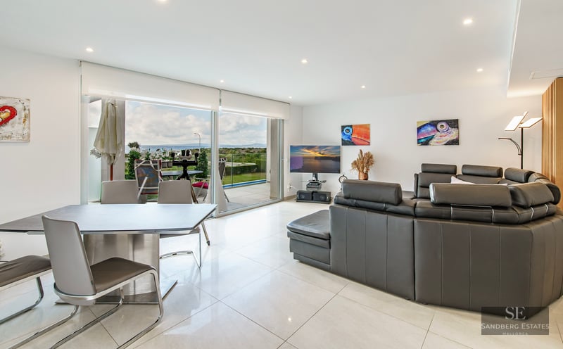 Modern living room with dark grey leather sofa, dining area, and large glass doors opening to a sea view.