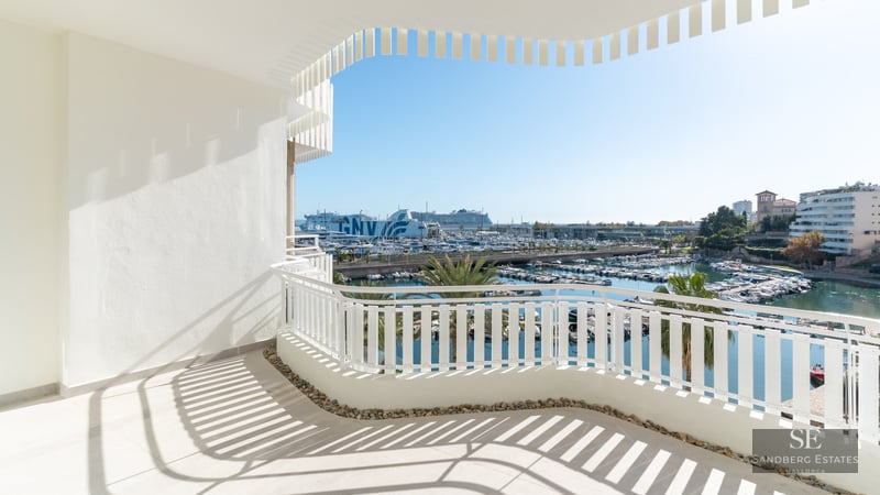 White curved balcony overlooking a sunny marina filled with boats and palm trees under a clear blue sky.