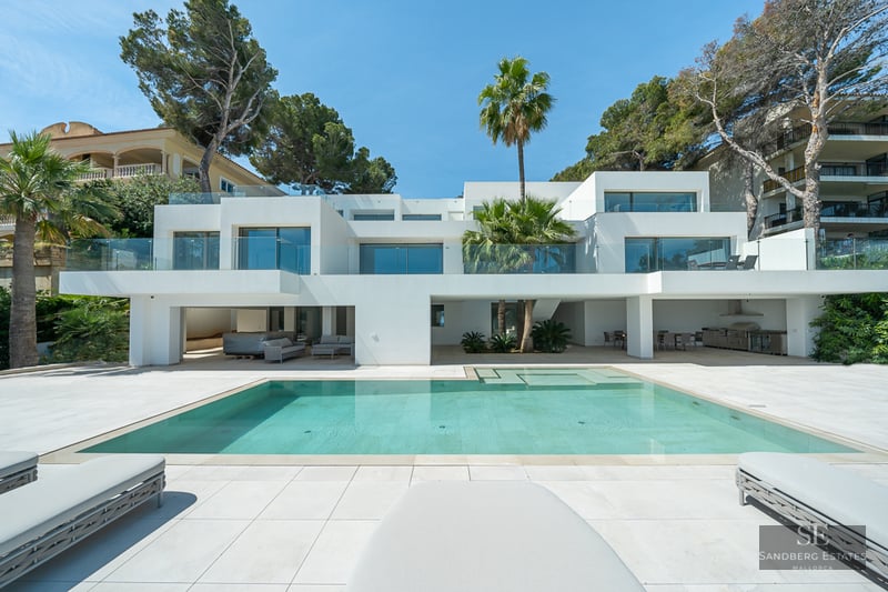 Modern white villa with a large turquoise pool, glass balconies, and palm trees under a clear blue sky.