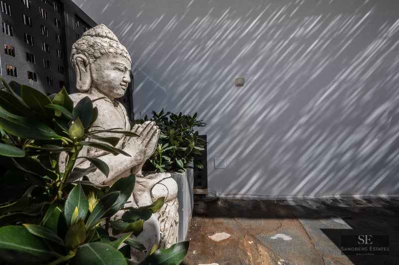 A stone Buddha statue tucked into green foliage on a terrace, with linear shadows falling on a white wall.