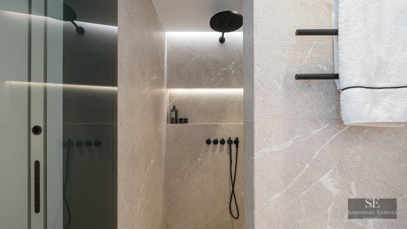 Walk-in shower with beige stone tiles, a matte black rain shower head, and an illuminated recessed niche on veined stone.