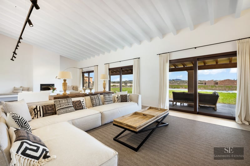 Bright living room with large L-shaped sofa, white wooden ceiling beams, and glass doors opening to a scenic terrace.