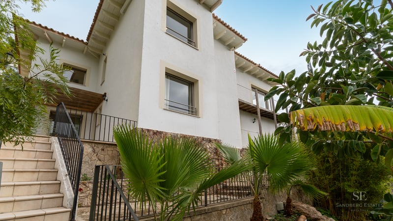 Three-story white villa facade with stone foundation, exterior stairs, and palm trees under a clear sky.