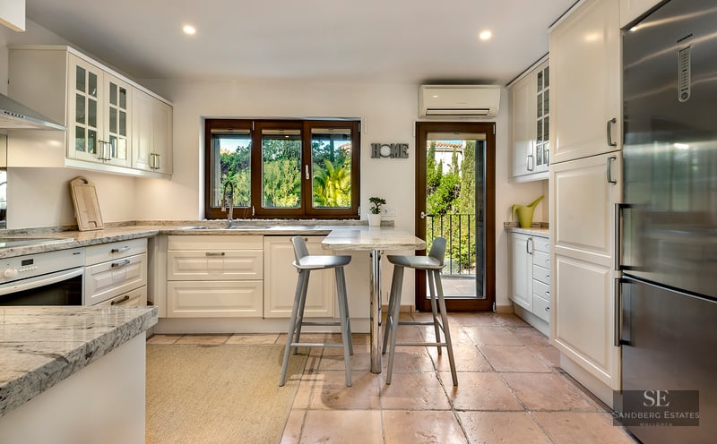 Bright kitchen with cream cabinets, granite counters, terracotta floors, breakfast bar, and window view of lush greenery.