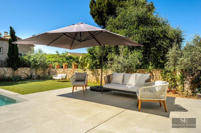 Sun-drenched terrace with modern grey sofa, armchairs, and large parasol next to a pool and lush garden.