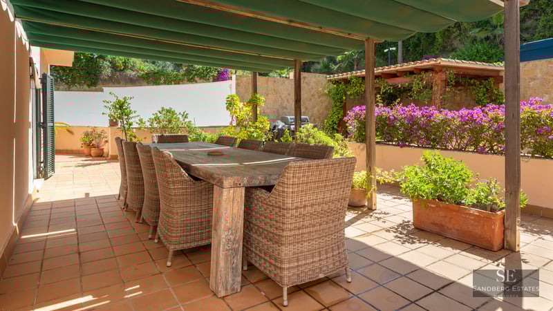 Large wooden dining table with wicker chairs on a sunny terracotta terrace under a green awning.
