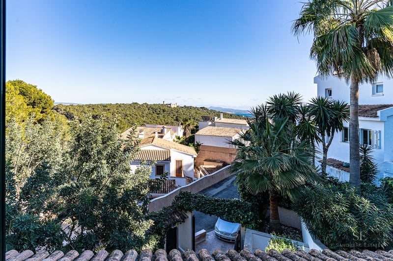 Elevated panoramic view showing white villas, lush greenery, a distant castle on a hill, and the sea under a blue sky.