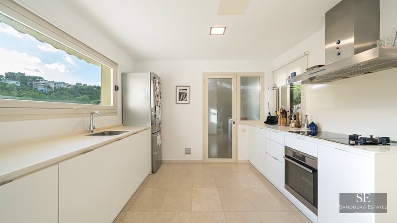 Bright kitchen with white cabinets, stainless steel fridge and oven, and a large window overlooking green trees.