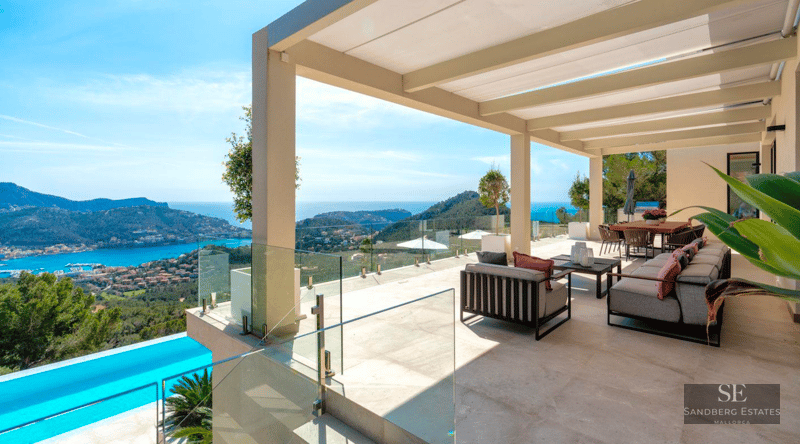 Large modern terrace with grey sofas, glass railings, and a stunning view of a blue bay and green hills.