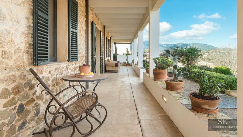 A long covered stone terrace with terracotta pots, green shutters, and a rocking chair overlooking mountains.