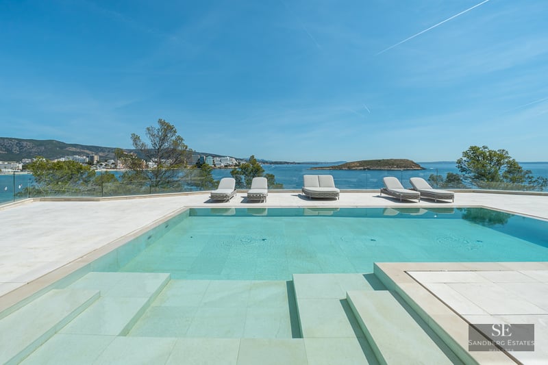 A turquoise swimming pool on a stone terrace with sun loungers overlooking the Mediterranean sea and a distant island.