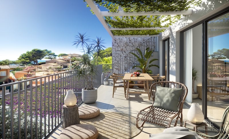 Sunny modern terrace with a wooden dining table, wicker lounge chair, and a plant-covered pergola overlooking a coastal town.