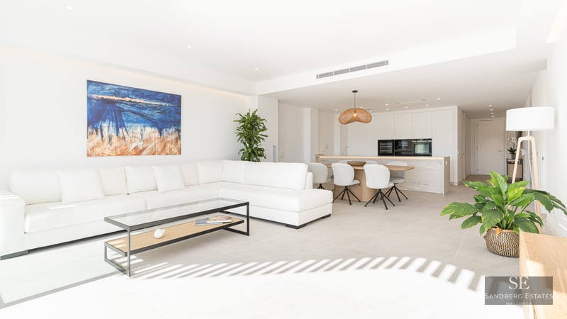 Bright living room with white L-shaped sofa, glass coffee table, abstract art, and integrated kitchen and dining space.