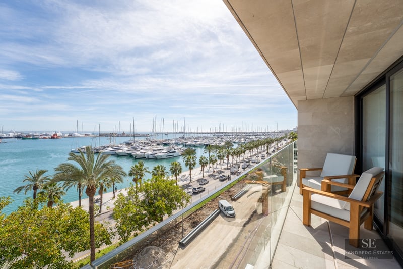 Modern balcony with wooden armchairs and glass railing overlooking a marina filled with yachts and palm trees.