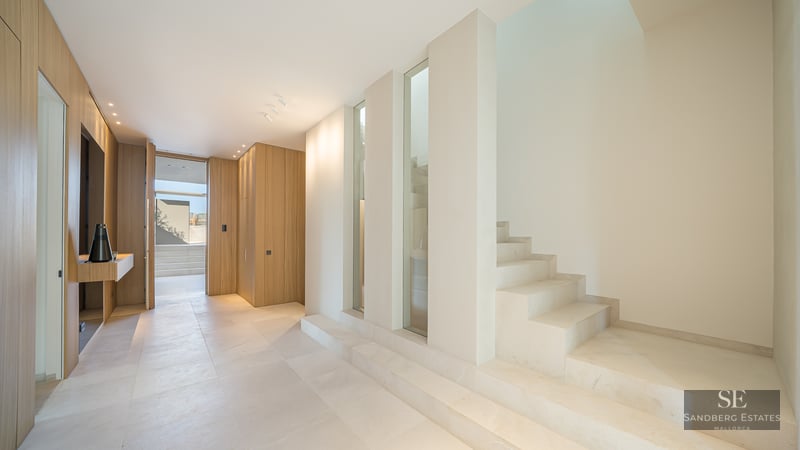 Modern hallway with cream stone floors, a minimalist stone staircase, and light oak wood paneling under bright light.