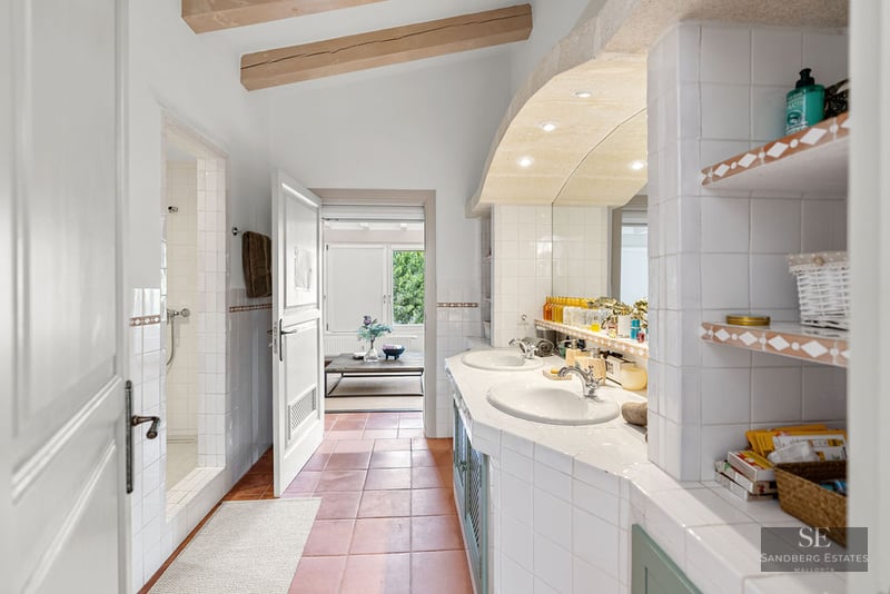 Spacious bathroom with terracotta floors, double white sinks, wooden ceiling beams, and a walk-in tiled shower.