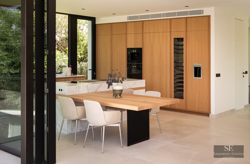 Modern kitchen with wood cabinets, marble island, integrated dining table, and large glass windows showing garden views.