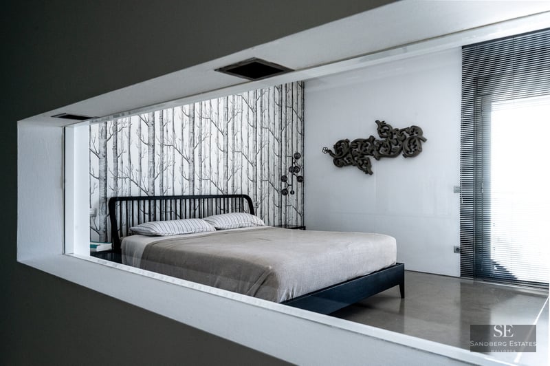 View of a modern bedroom through a wall opening showing a bed with tree-patterned wallpaper and unique wall art.