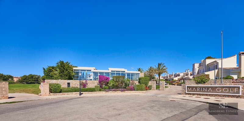 Stone entrance with "Marina Golf" sign under a clear blue sky and Mediterranean landscaping.