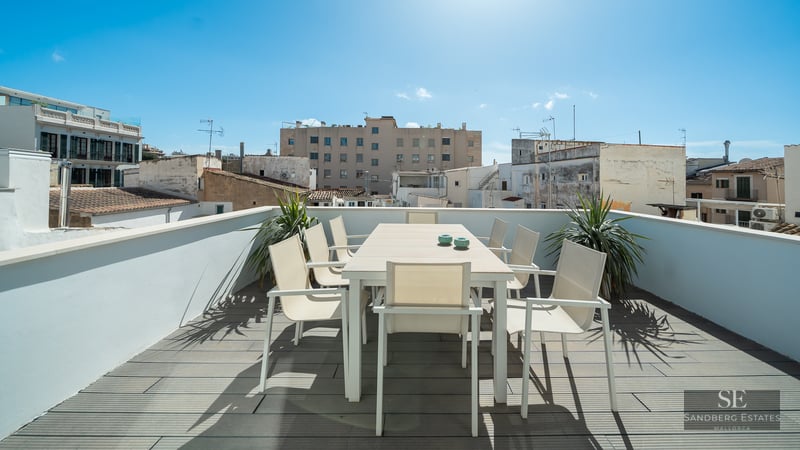 Modern rooftop terrace with white dining set on grey wooden decking overlooking city buildings under a blue sky.