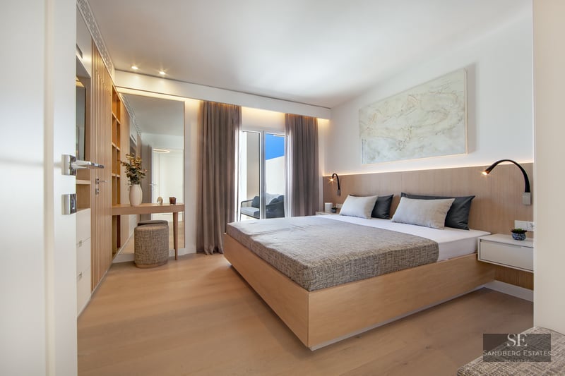 Modern master bedroom with a large wooden bed, integrated lighting, wood paneling, and access to a sunny balcony.