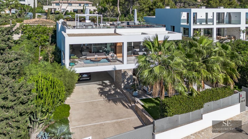 Vue architecturale d'une villa moderne blanche avec terrasses, palmiers et murs en pierre sous une lumière vive.