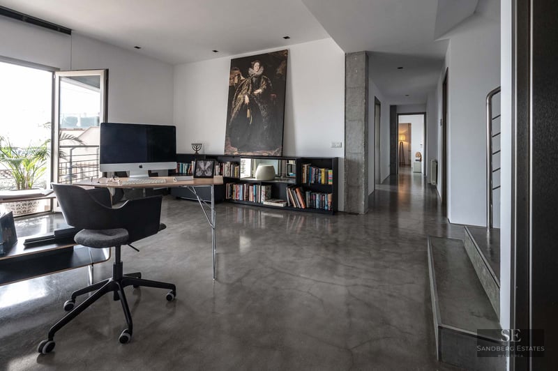 A minimalist home office featuring a desk, iMac, large portrait art, and polished concrete floors leading to a hallway.