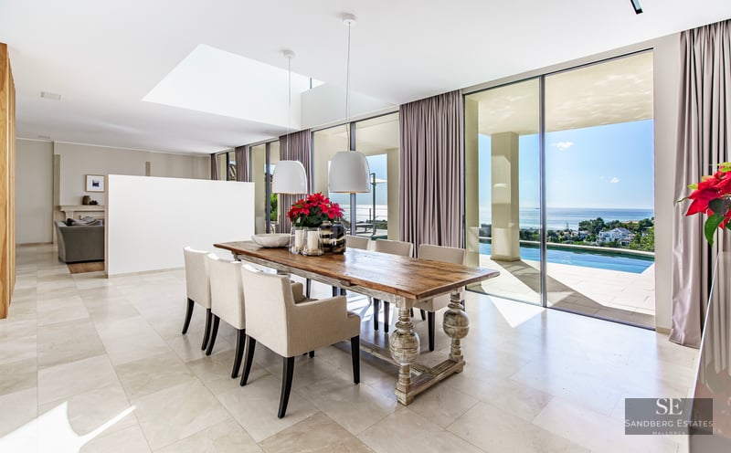 Spacious dining room with a large wooden table, cream chairs, and floor-to-ceiling windows overlooking a sea and pool.