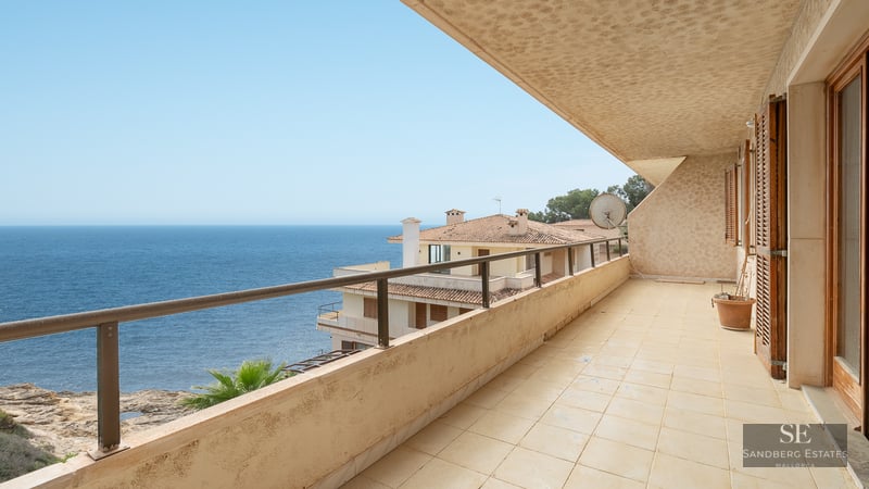 A wide tiled balcony with a metal railing overlooking the blue ocean and nearby Mediterranean buildings.