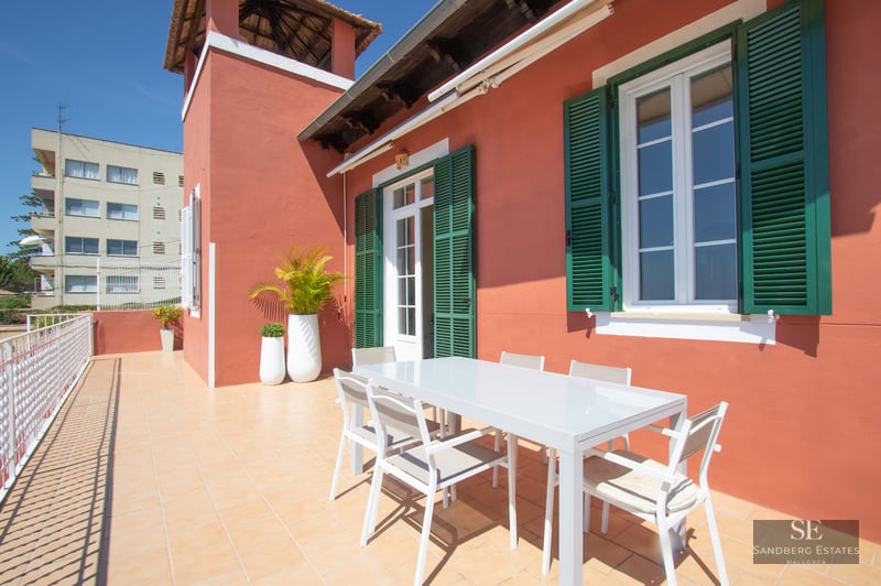 A sunlit terrace with terracotta walls, green shutters, and a white modern dining set under a clear blue sky.