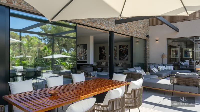 Modern terrace featuring a large wooden dining table, grey sofas, and floor-to-ceiling glass doors to the living room.