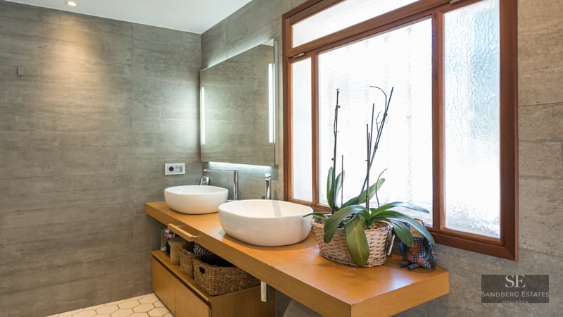 Contemporary bathroom with two white vessel sinks on a wooden vanity, grey tiled walls, and a large backlit mirror.