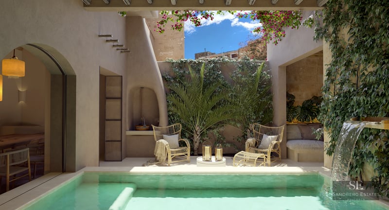 Sunlit courtyard with turquoise plunge pool, waterfall, wicker chairs, and lush greenery under a blue sky.