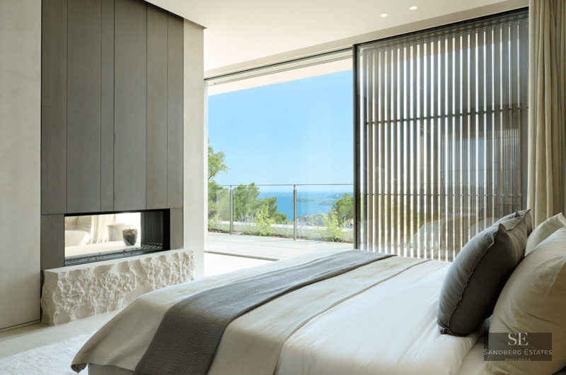 Luxury bedroom featuring floor-to-ceiling windows overlooking the ocean and a modern fireplace with a rough stone base.
